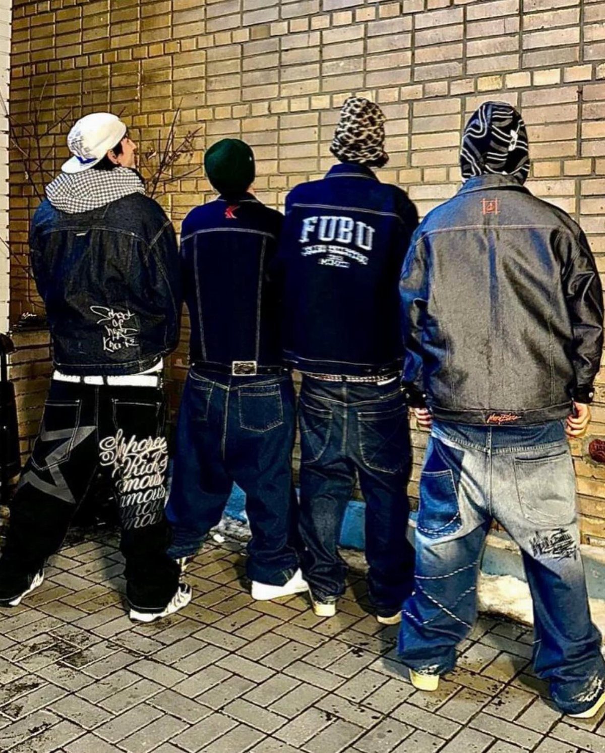 Four people in baggy 2000s hip hop fashion standing against a brick wall.