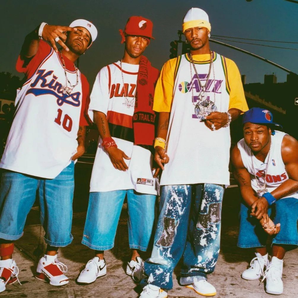 Four men posing in oversized basketball jerseys and baggy streetwear on an urban rooftop.