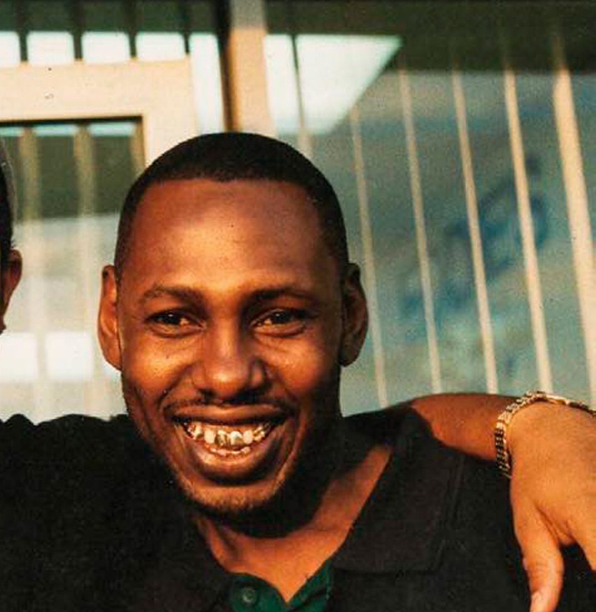 A smiling man with gold teeth and a gold chain bracelet posing outdoors.