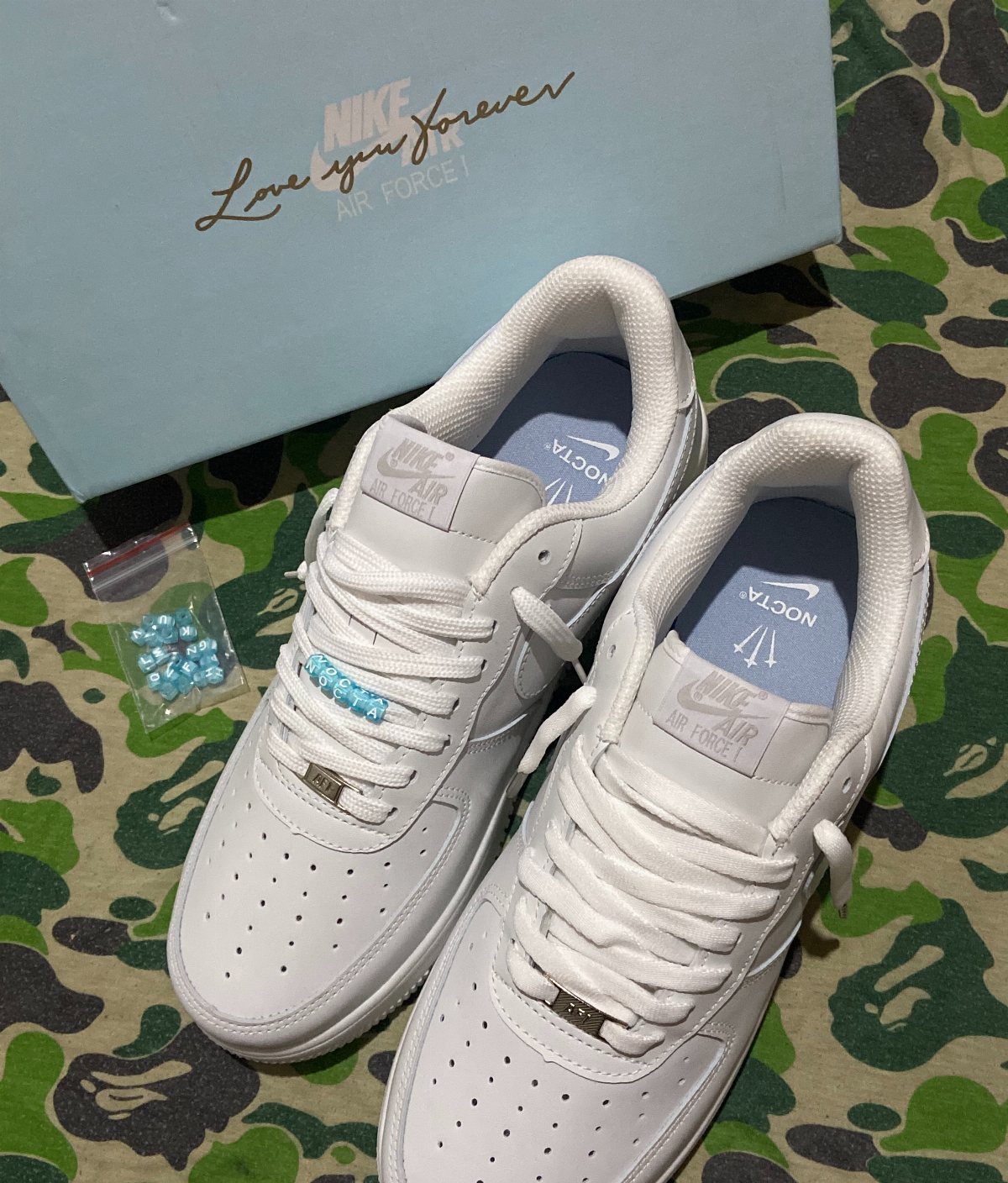 White Nike Air Force 1 sneakers with blue charms resting on camouflage fabric near a shoebox.
