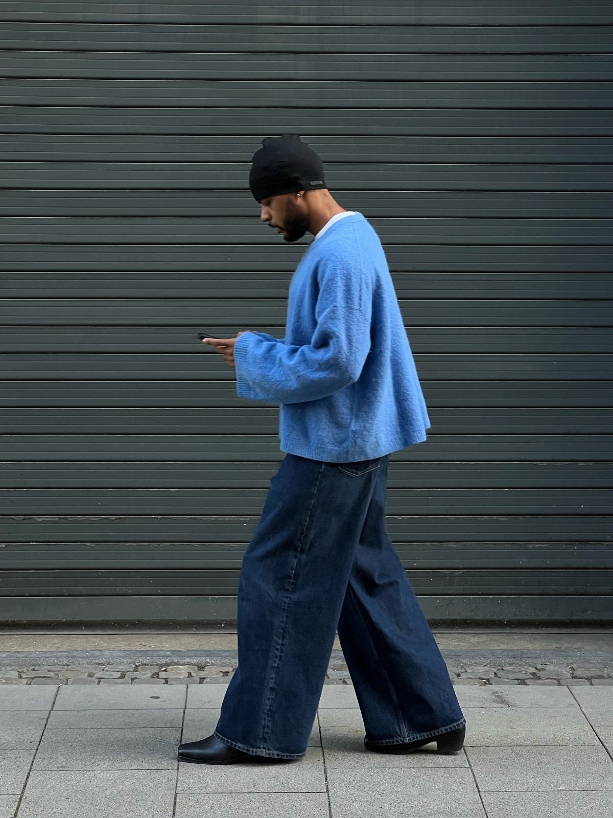 A man in a blue knit sweater and wide-leg denim jeans walking past a grey wall.