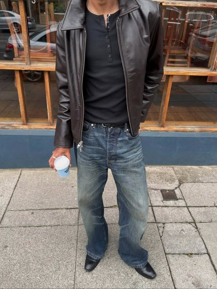 A man wearing a leather jacket, black henley, and blue bootcut denim jeans standing outdoors.