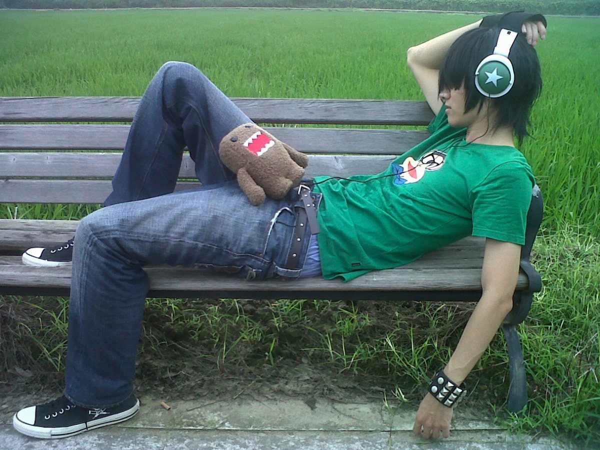 A young man with emo hair reclining on a wooden bench in a grassy field.