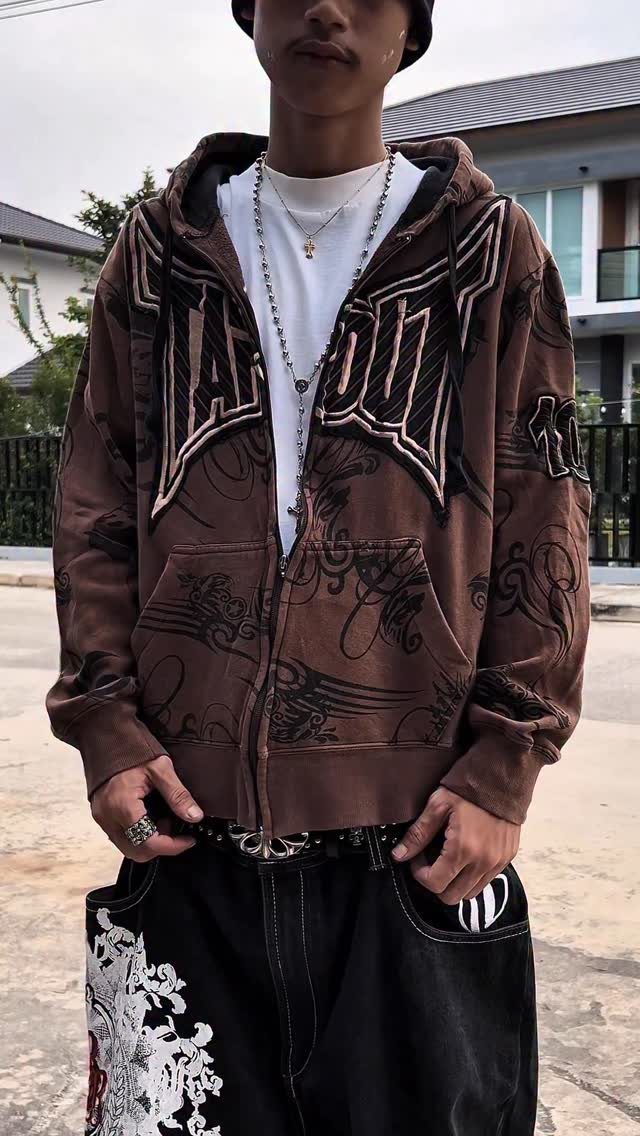 A person wearing a brown graphic zip-up hoodie, silver necklaces, and black patterned baggy pants.