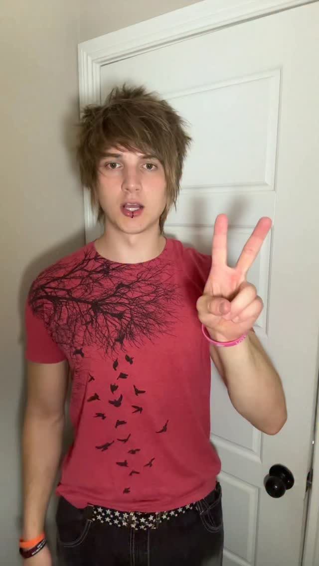 Young man with shaggy hair wearing a graphic red t-shirt and studded belt making a peace sign.