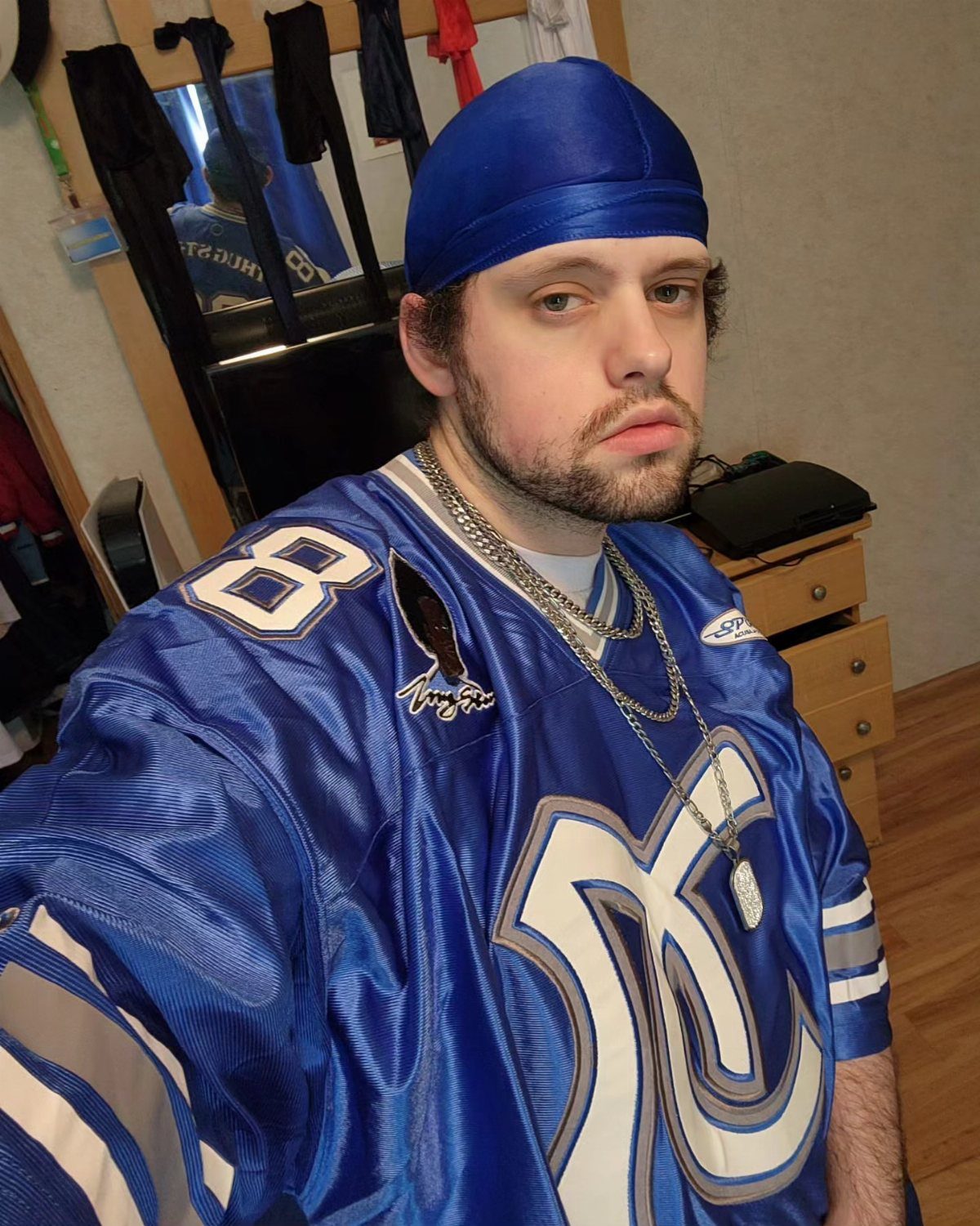 A man wearing a blue durag, a blue sports jersey, and silver chain necklaces.