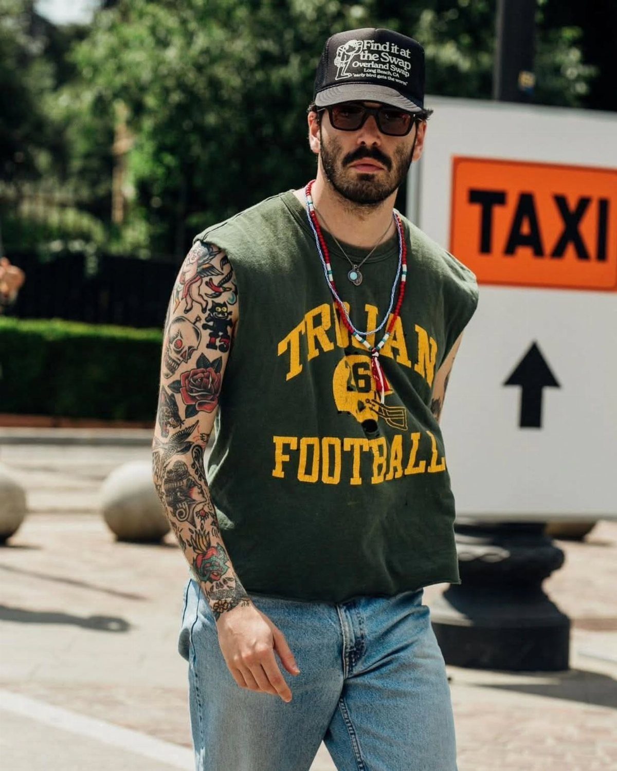 A tattooed man wearing a green sleeveless football shirt, denim jeans, and a black cap.