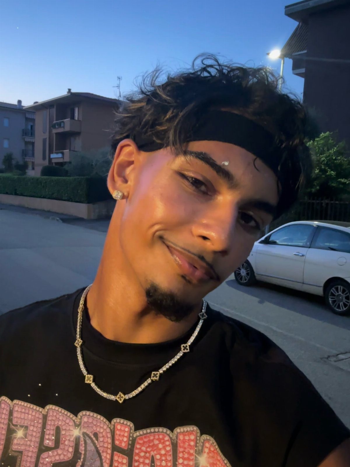 A smiling young man wearing a black headband and a silver chain necklace outdoors.
