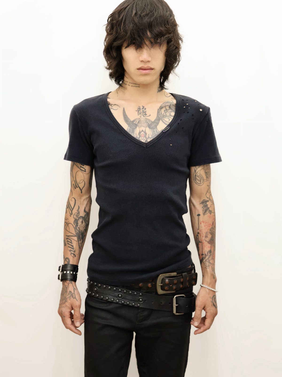 A man with tattoos wearing a tight navy blue V-neck t-shirt and a studded belt.