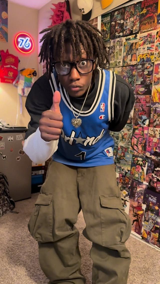 A young man wearing a blue basketball jersey and olive green cargo pants giving a thumbs up.