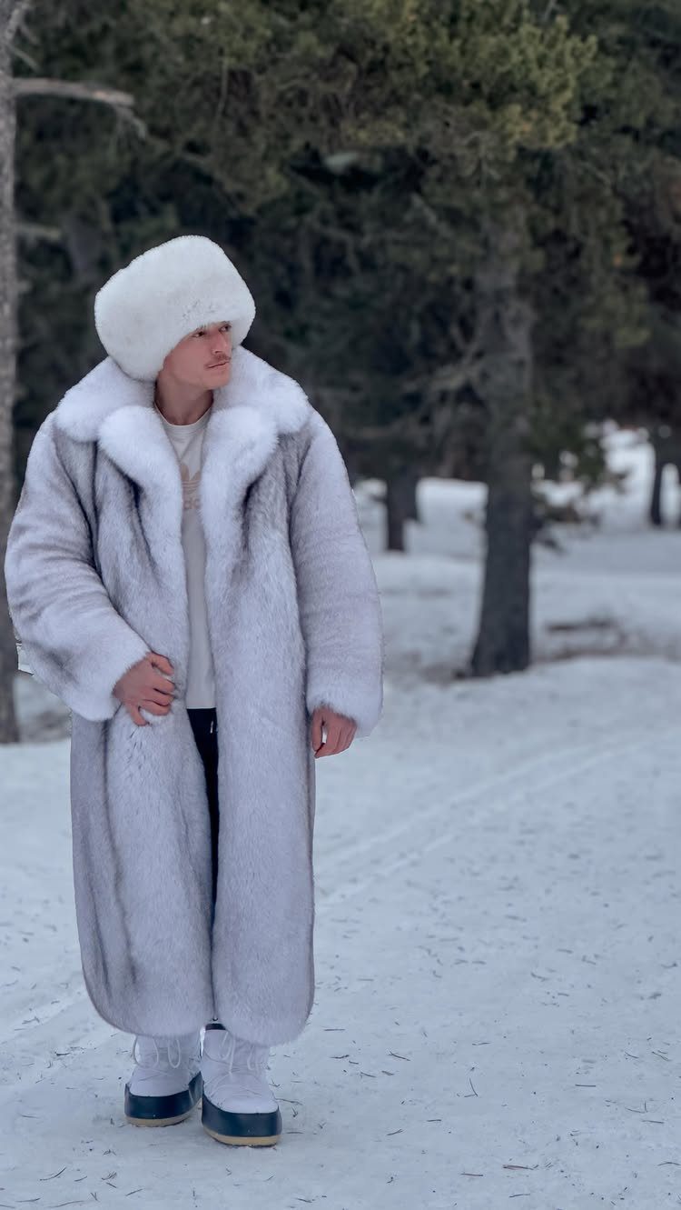 A man in a long grey fur coat and white hat standing in the snow.