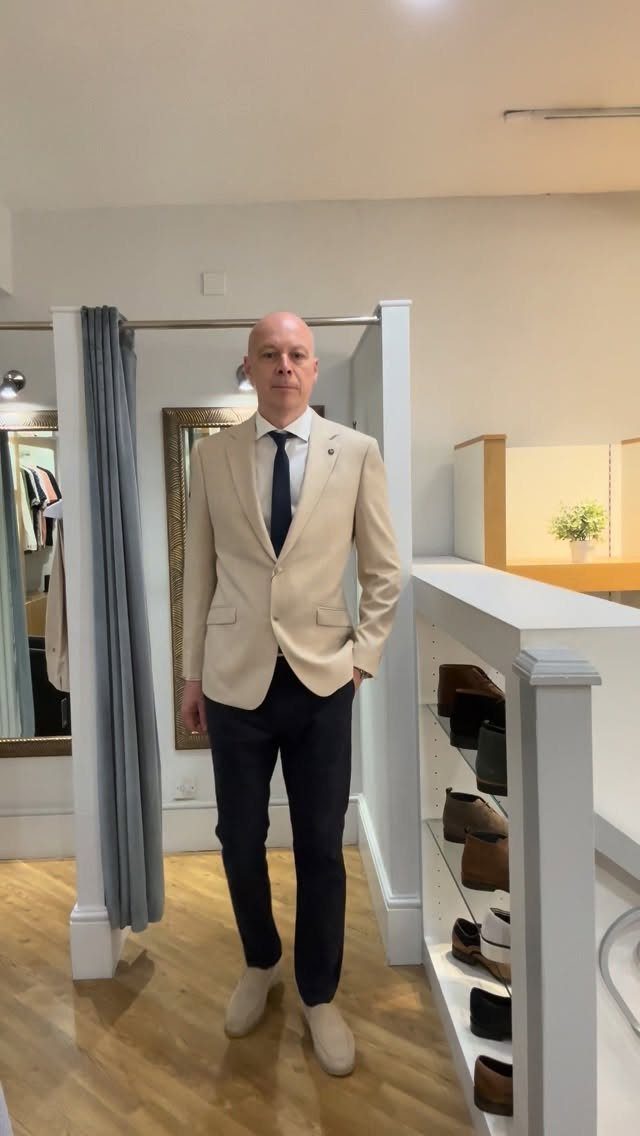 A man posing in a beige blazer, white shirt, dark tie, and navy trousers.