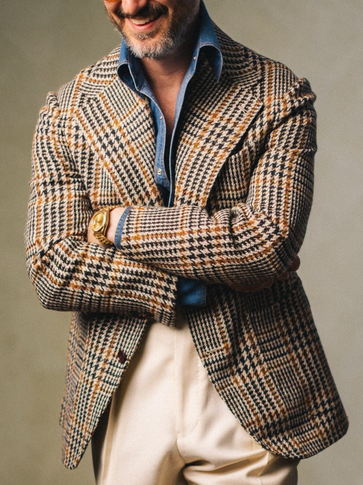 A smiling man in a houndstooth blazer, denim shirt, and cream trousers posing with arms crossed.