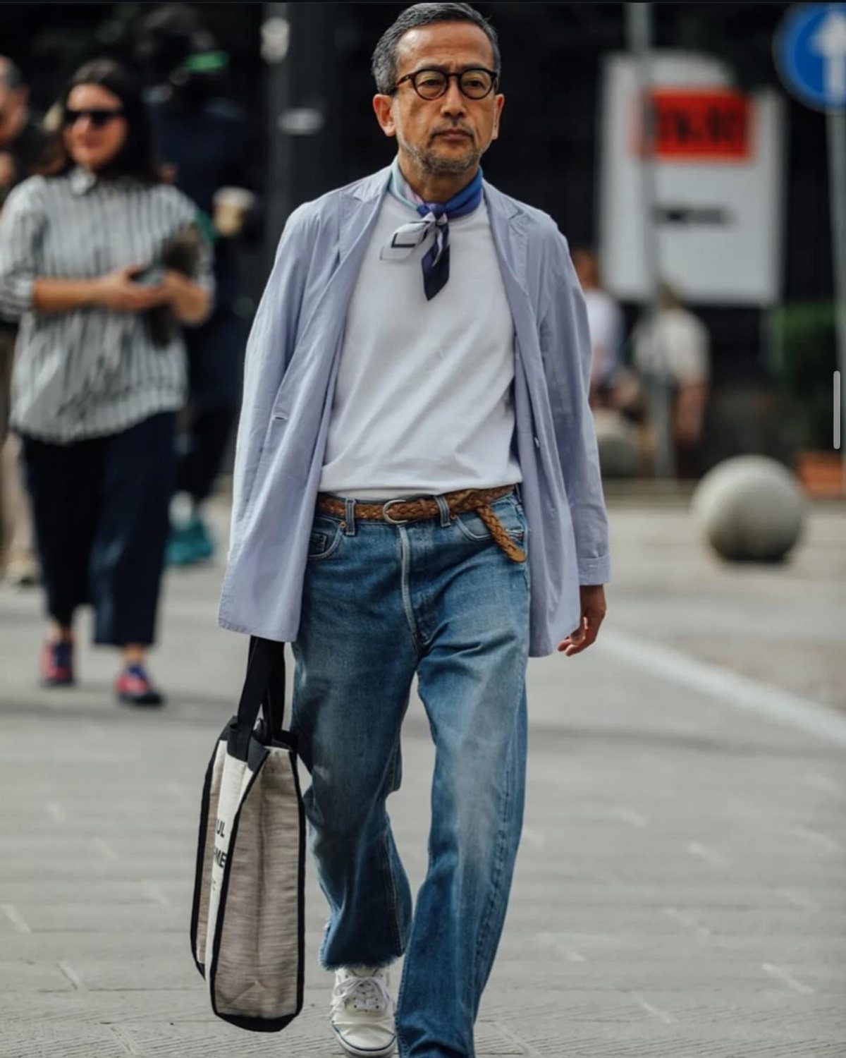 Man walking in a light blue blazer, white t-shirt, blue jeans, and a patterned neck scarf.