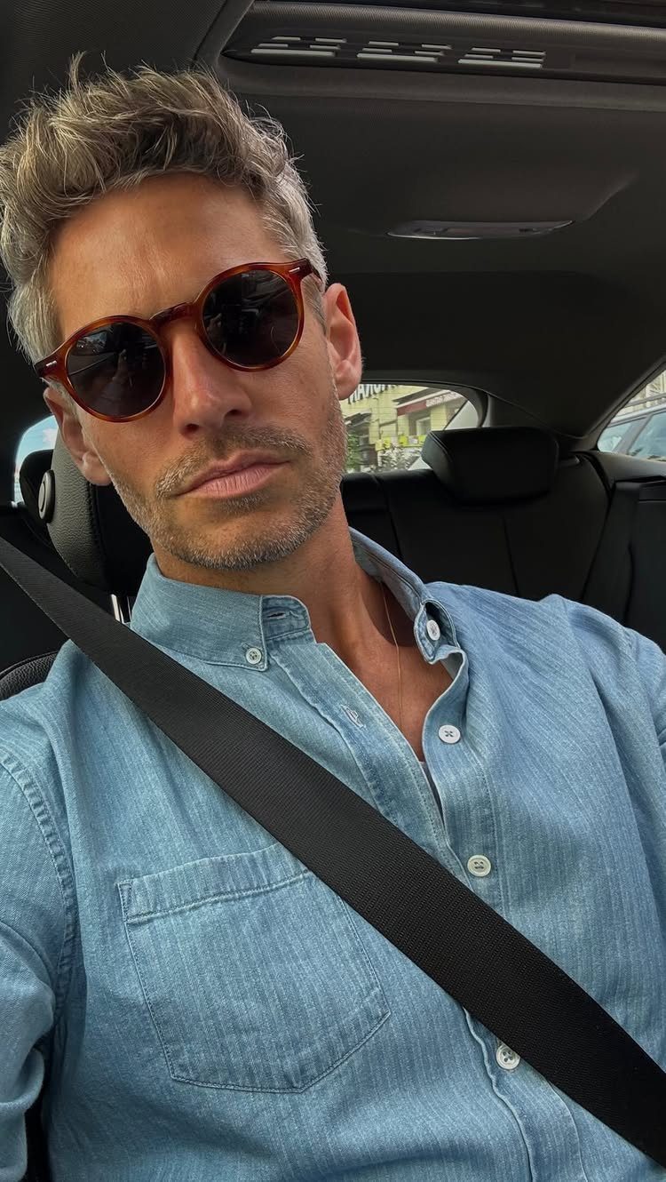 A man with grey hair wearing sunglasses and a blue denim shirt sitting in a car.