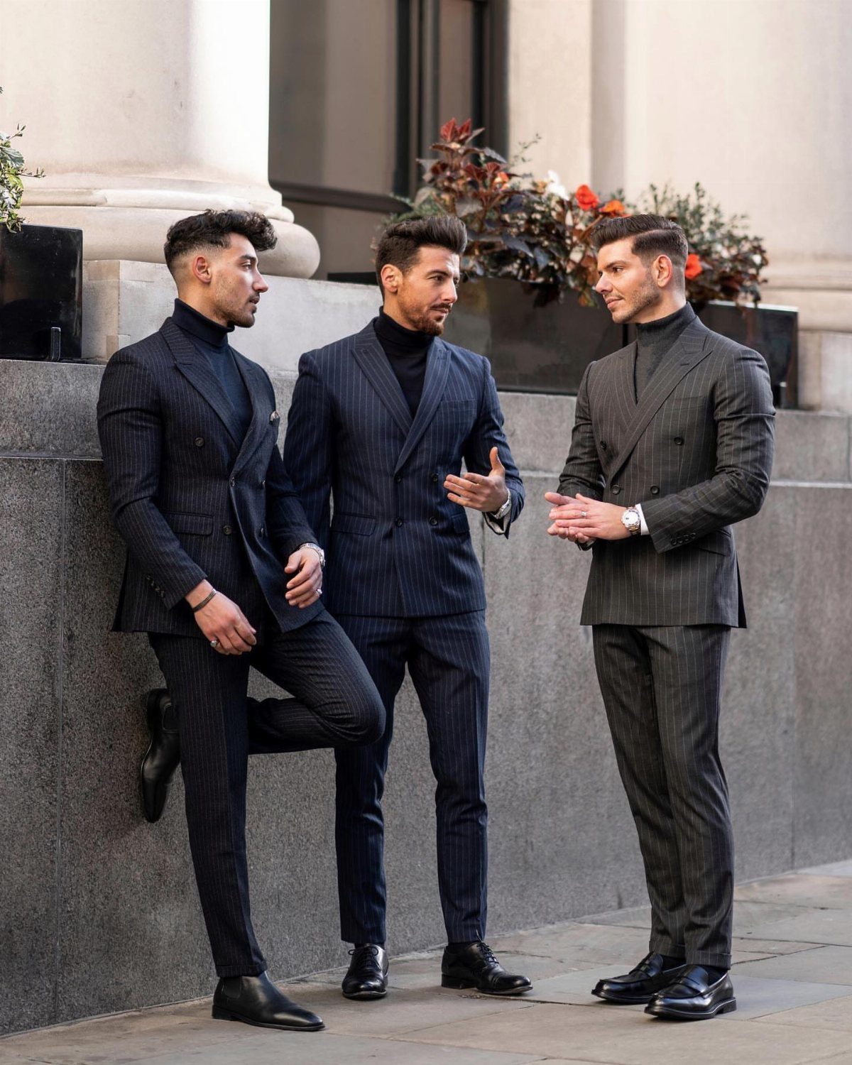 Three men in stylish pinstripe suits and turtlenecks standing and talking outdoors.