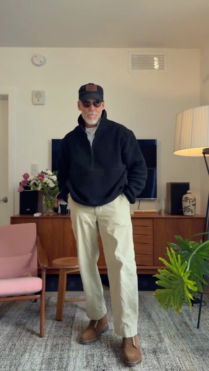 A man wearing a black fleece, beige pants, and a baseball cap posing indoors.