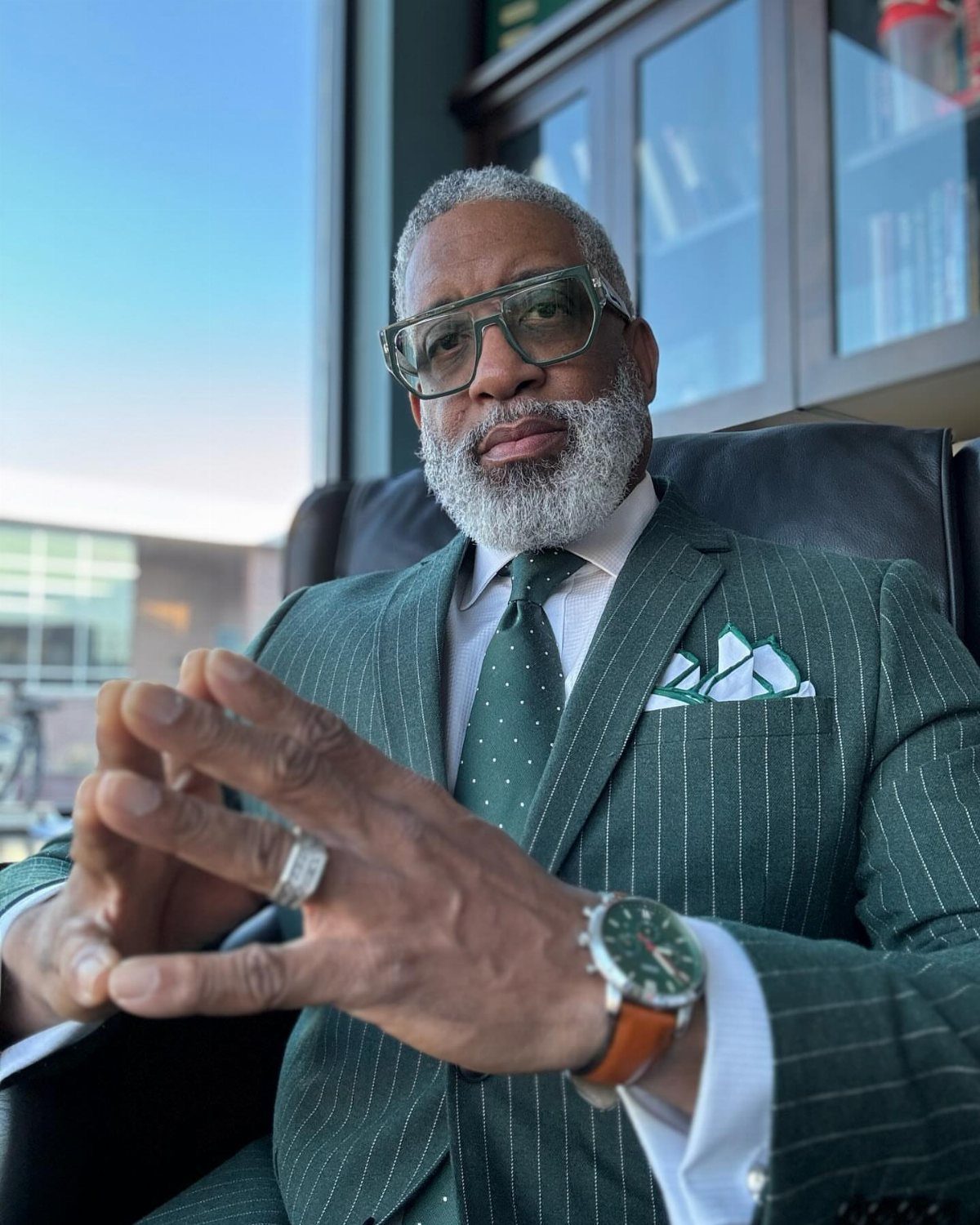 A mature man with a grey beard wearing a tailored green pinstripe suit and glasses.