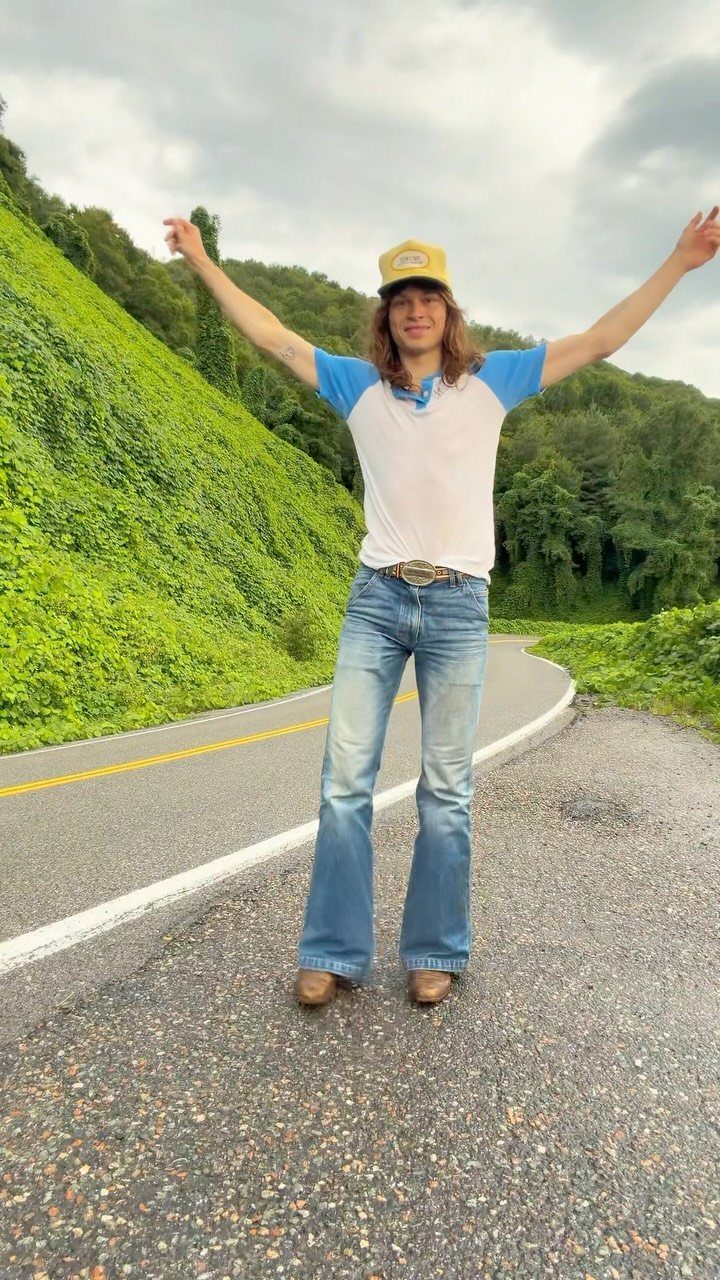 A person wearing denim bell-bottom jeans and a ringer tee stands on a road.