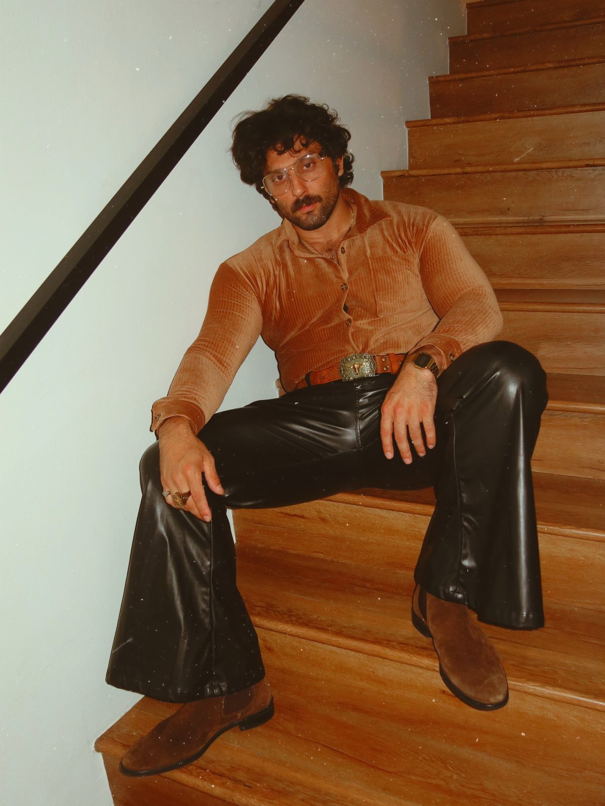 Man in corduroy shirt and black flared trousers sitting on wooden stairs.