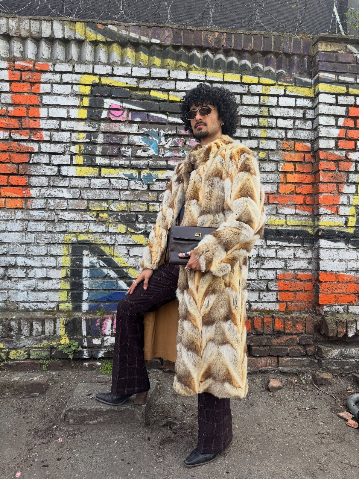 A man in a long faux fur coat and sunglasses posing against a graffiti wall.