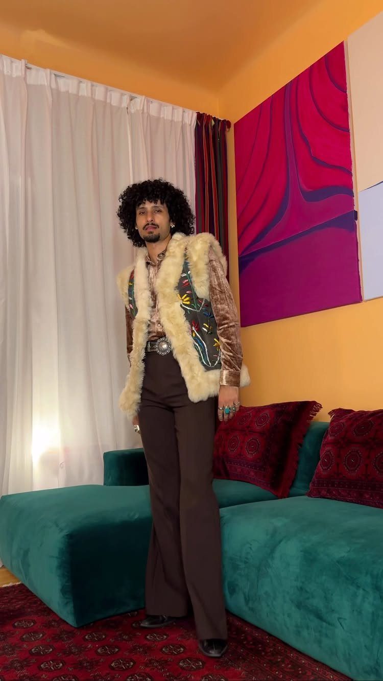 A man posing in a fur vest and flared brown trousers in a colorful room.
