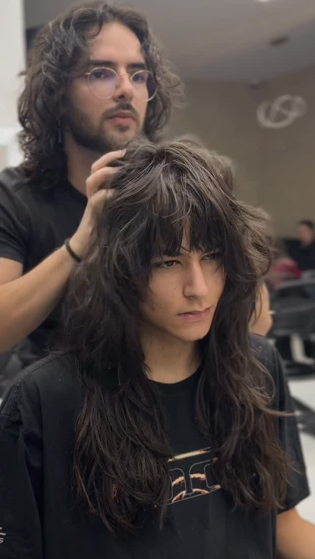 A person with voluminous, dark, layered shaggy hair being styled by a man in a salon.