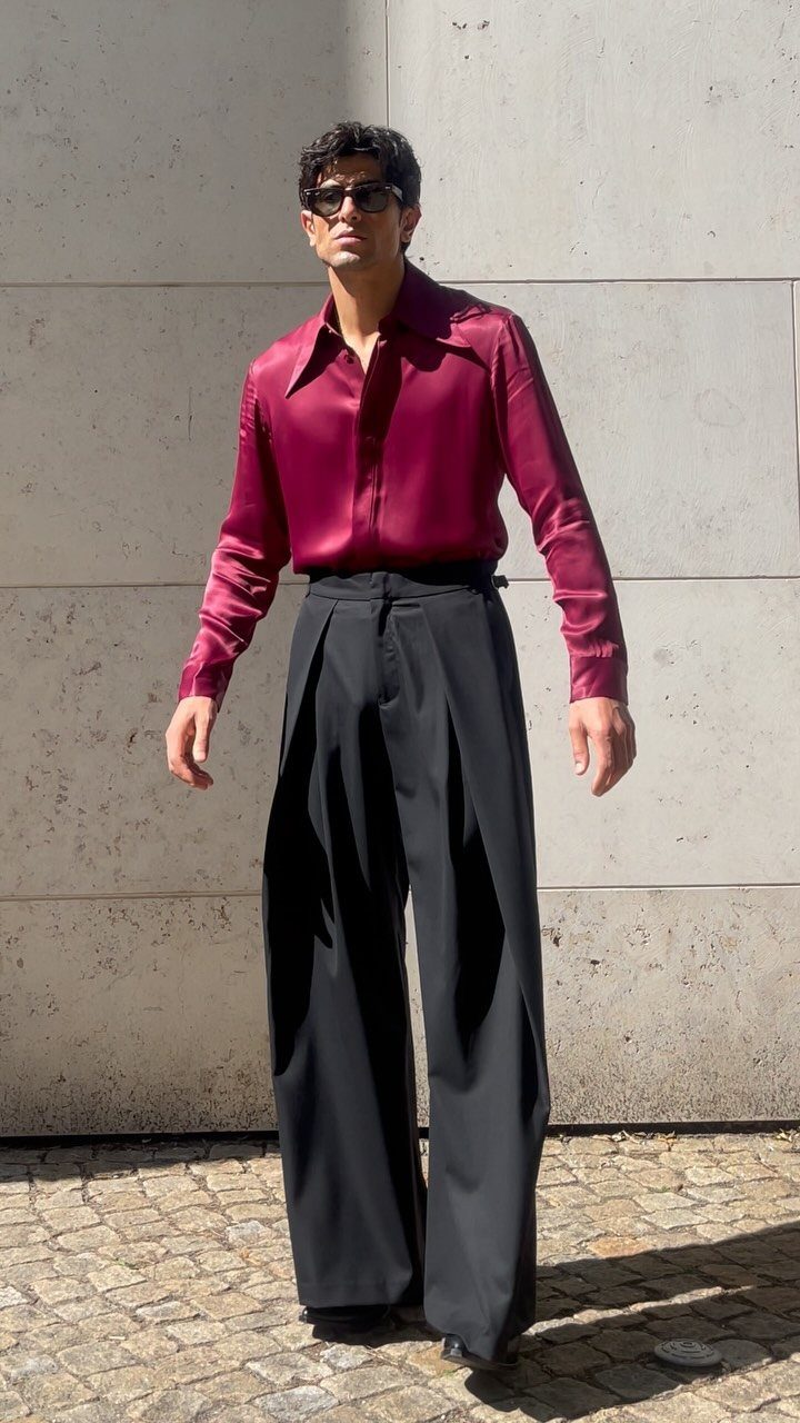 Man in a burgundy silk shirt and wide-leg black trousers standing against a stone wall.