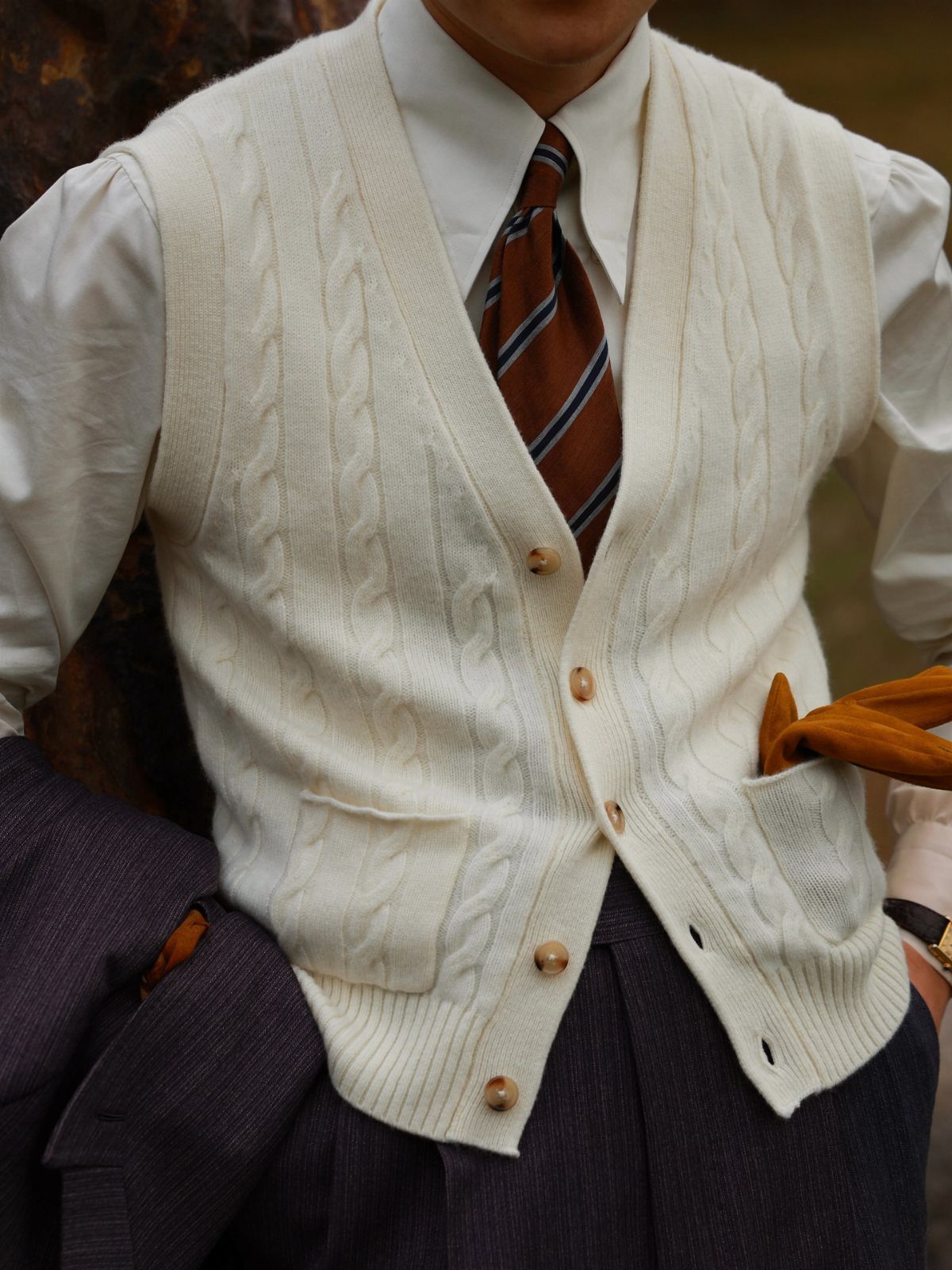 A person wearing a cream cable-knit sweater vest, white shirt, and striped tie.