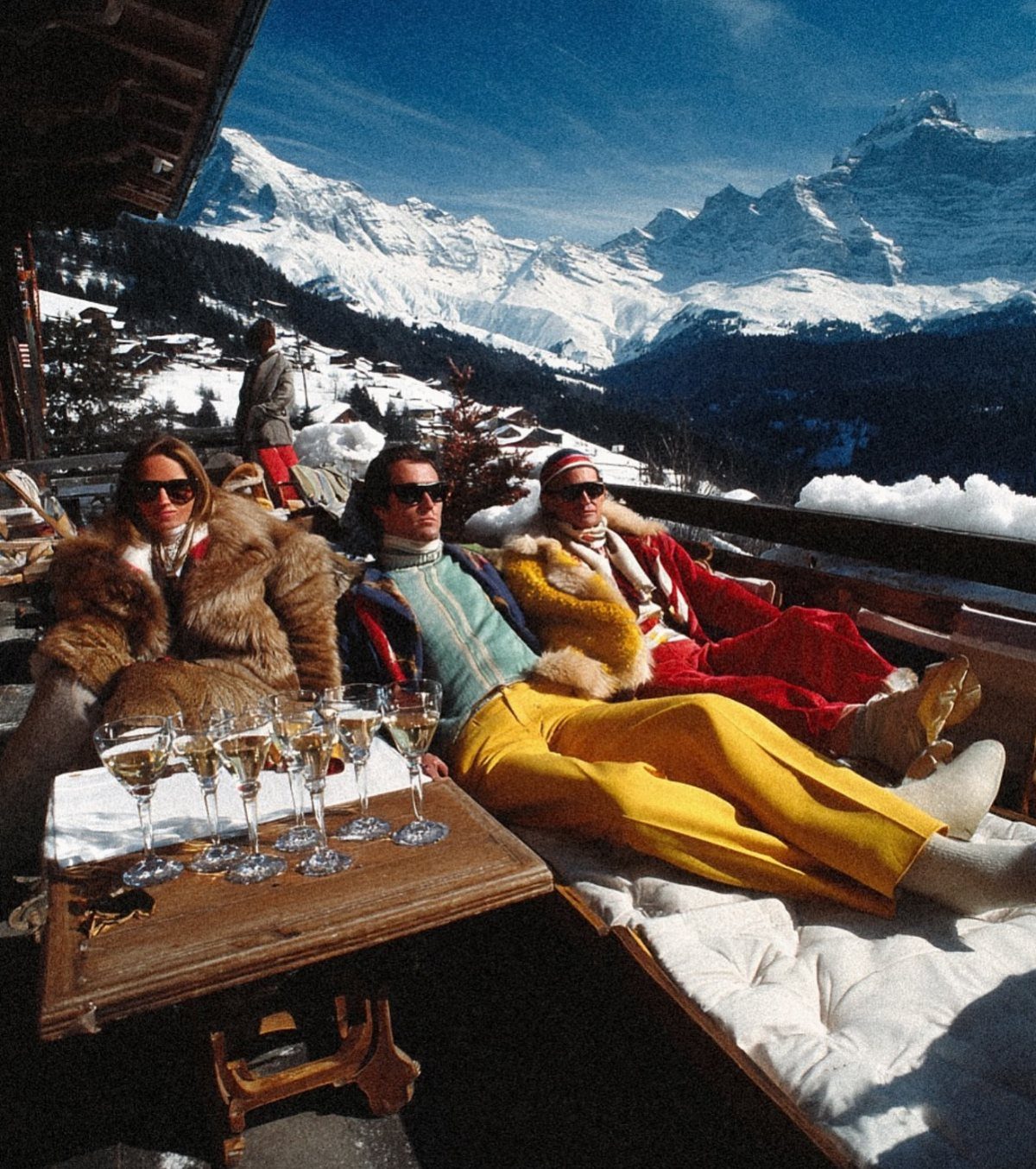 People in fashionable winter attire lounging on a terrace overlooking snowy mountain peaks.