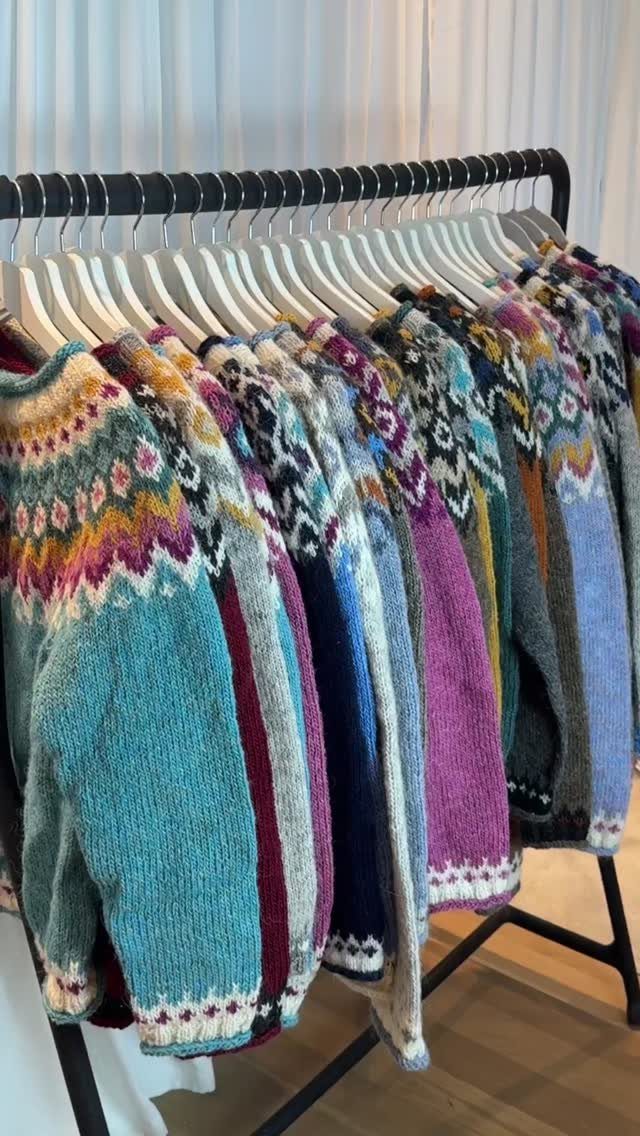 Colorful patterned knit sweaters hanging on a black clothing rack against a white curtain background.