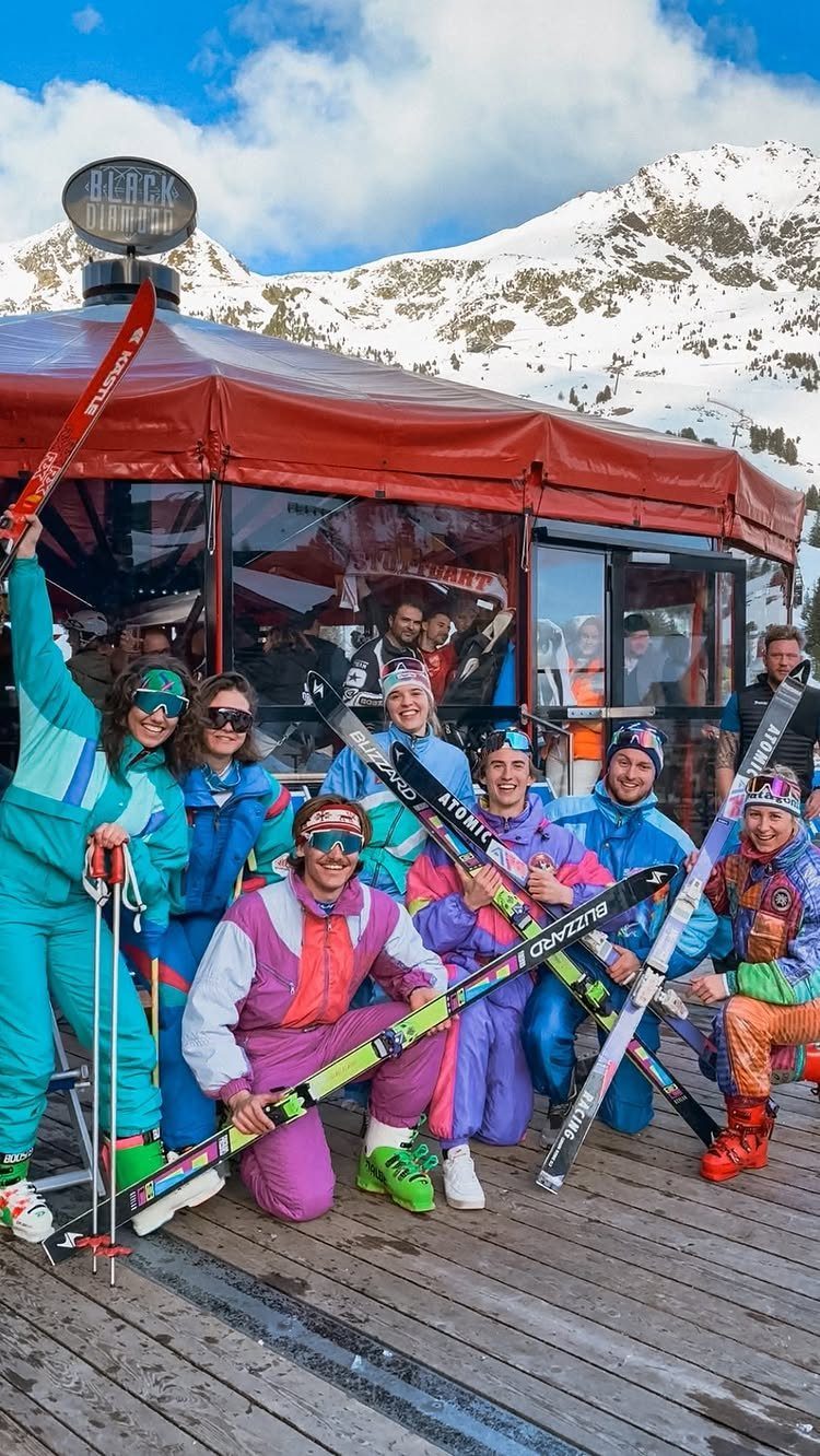 Group of people in colorful vintage ski suits posing with skis against snowy mountains.