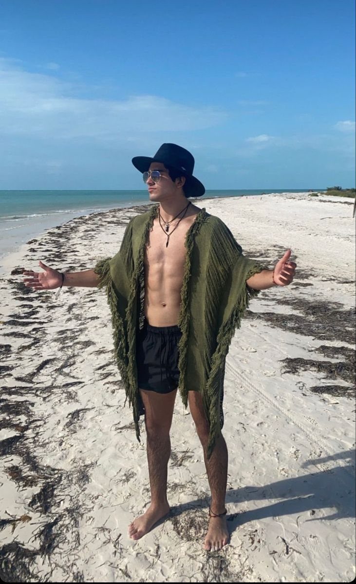 A man wearing a green fringed beach cover-up and black hat standing on a sandy beach.