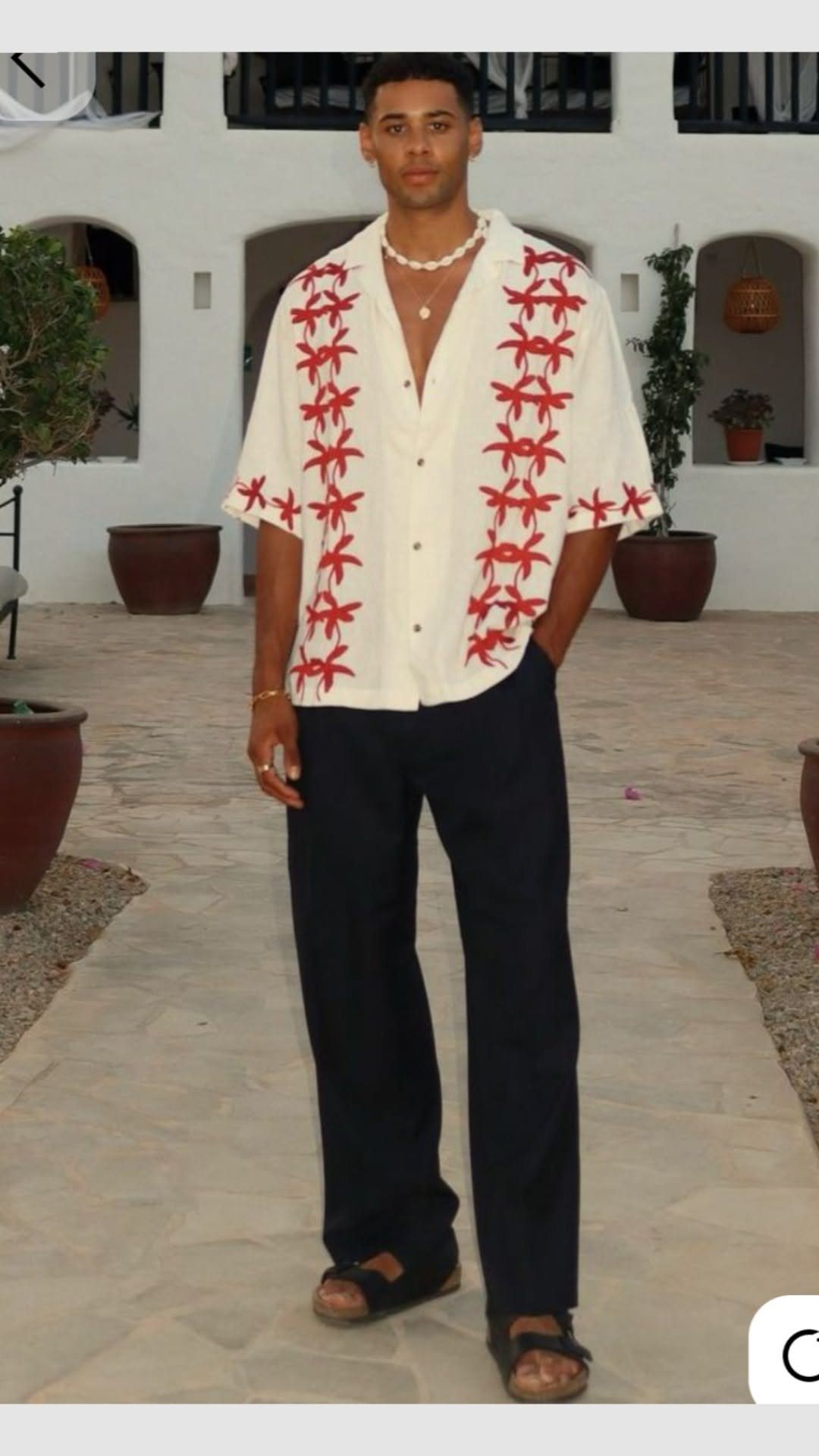 A man wearing a white embroidered shirt and dark trousers standing in a courtyard.
