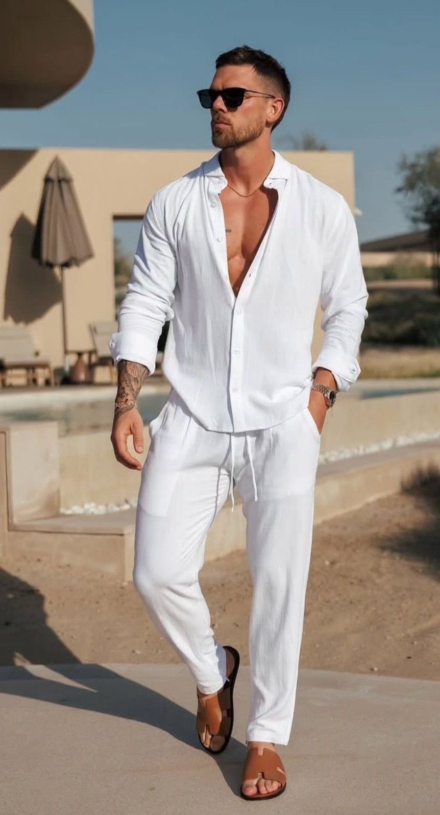 Man in a white linen shirt and matching trousers walking outdoors near a swimming pool.
