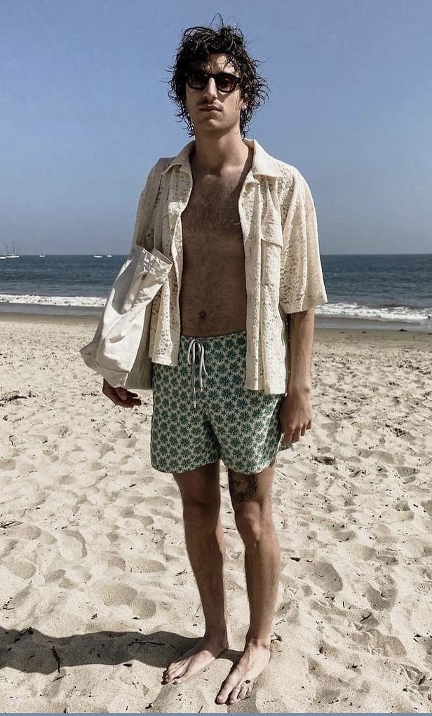 A man in an open beige shirt and patterned green swim shorts standing on a sandy beach.