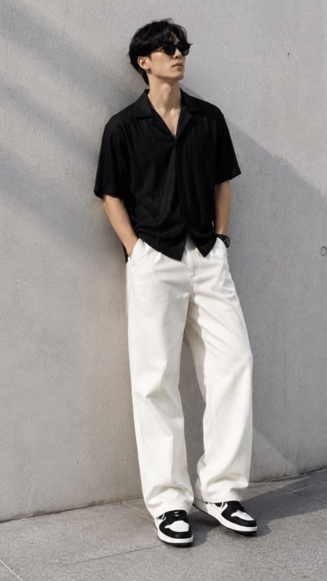 Man in black short-sleeved shirt and wide white trousers leaning against a wall.