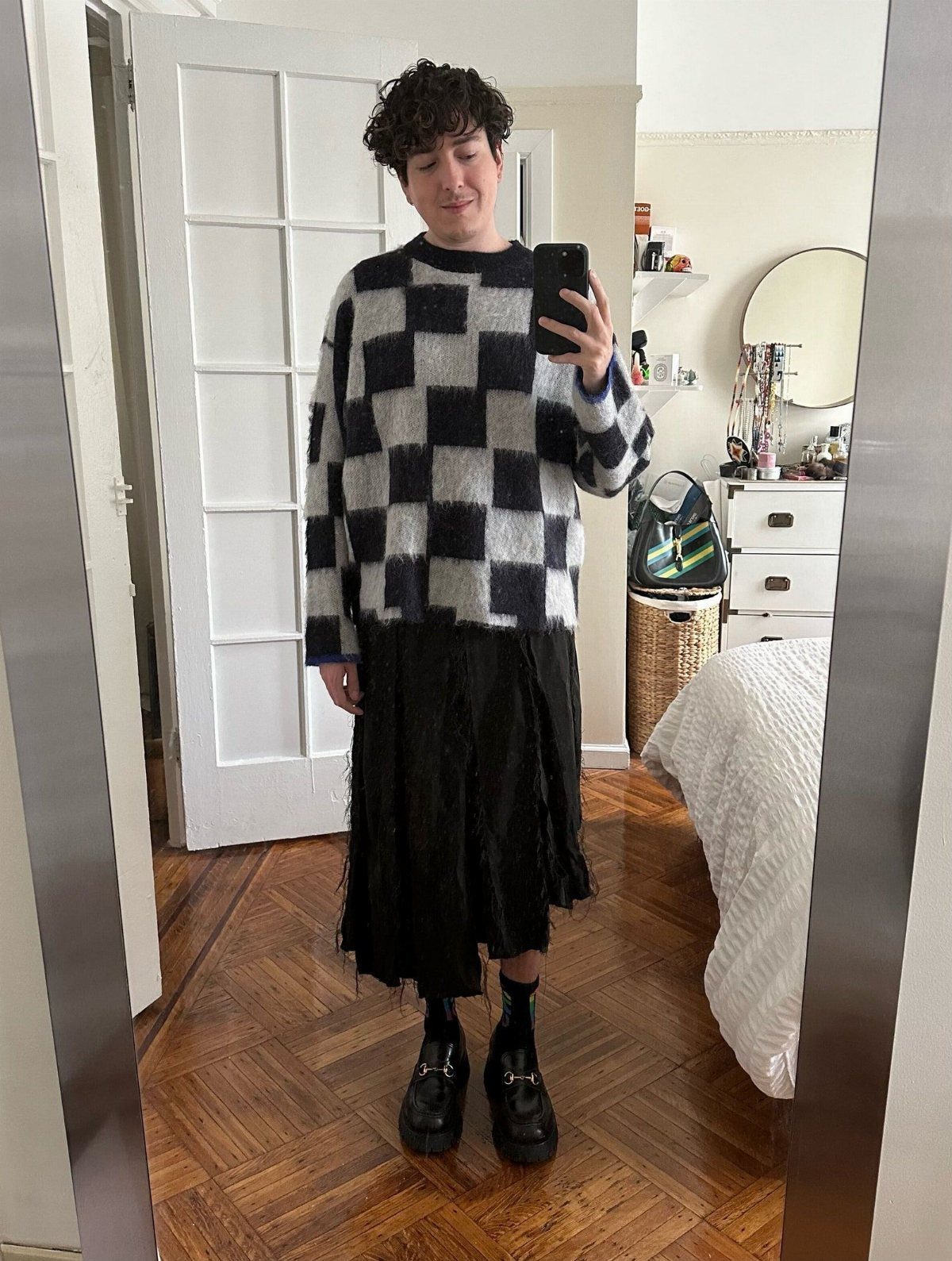 Person wearing a checkered sweater, black skirt, and black loafers standing in a room.