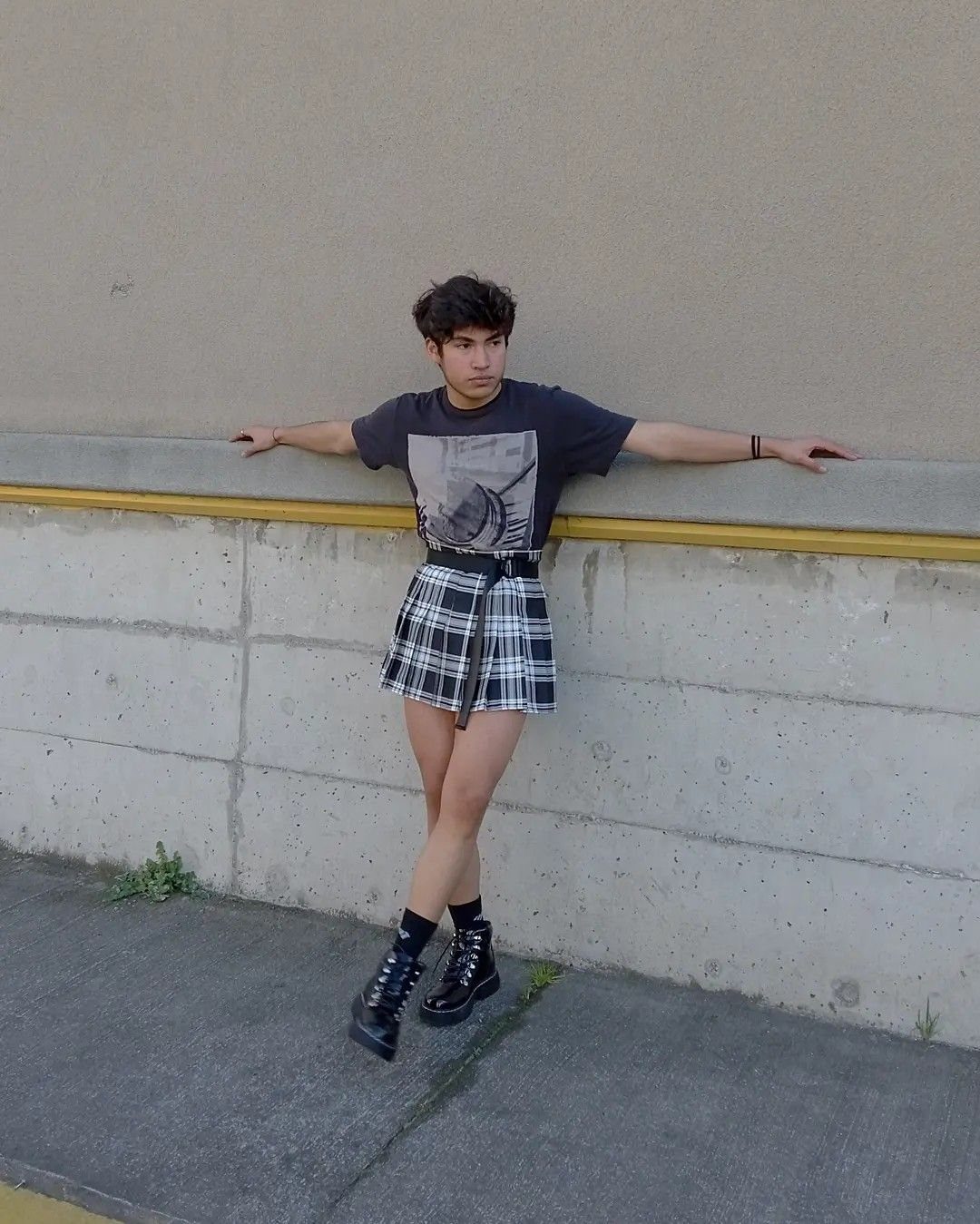 A person wearing a black graphic tee, plaid skirt, and black boots leaning against a wall.