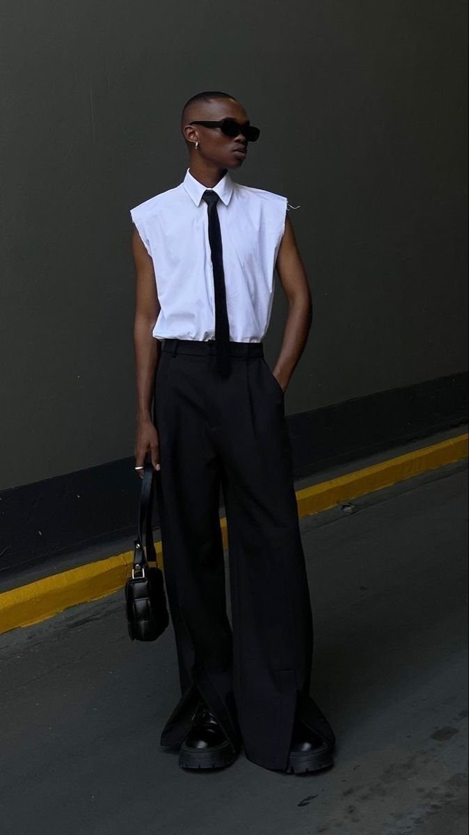 A person wearing a white sleeveless shirt, black tie, and wide-leg black trousers standing outdoors.