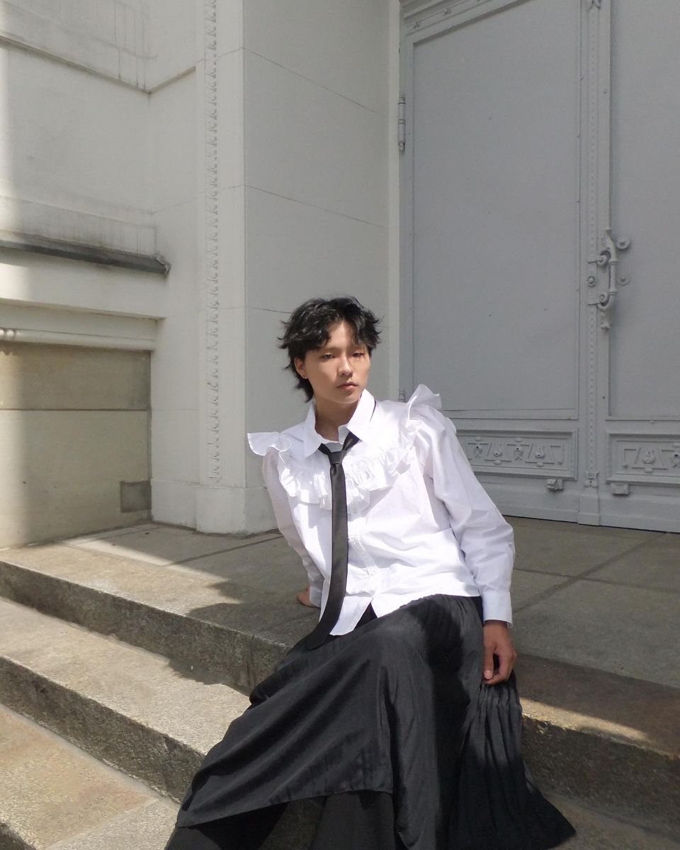 A person wearing a white ruffled shirt, black tie, and long black skirt sitting on steps.