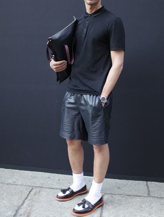 A man wearing a black polo shirt, black leather shorts, white socks, and loafers.