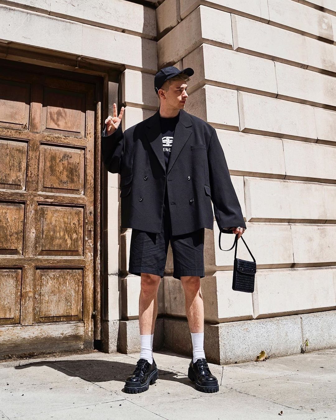 A person wearing an oversized black blazer, black shorts, and black shoes standing by a stone wall.
