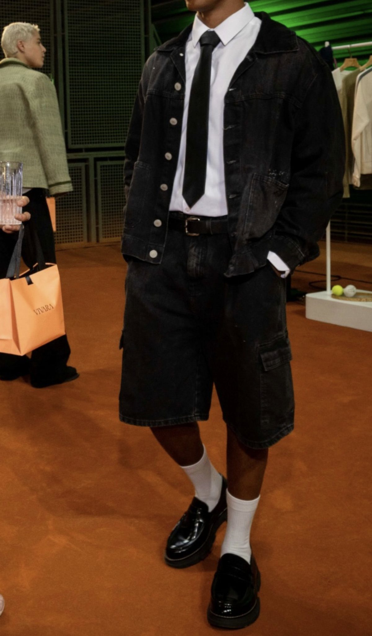A person wearing a black denim jacket, white shirt, black tie, and black denim shorts.