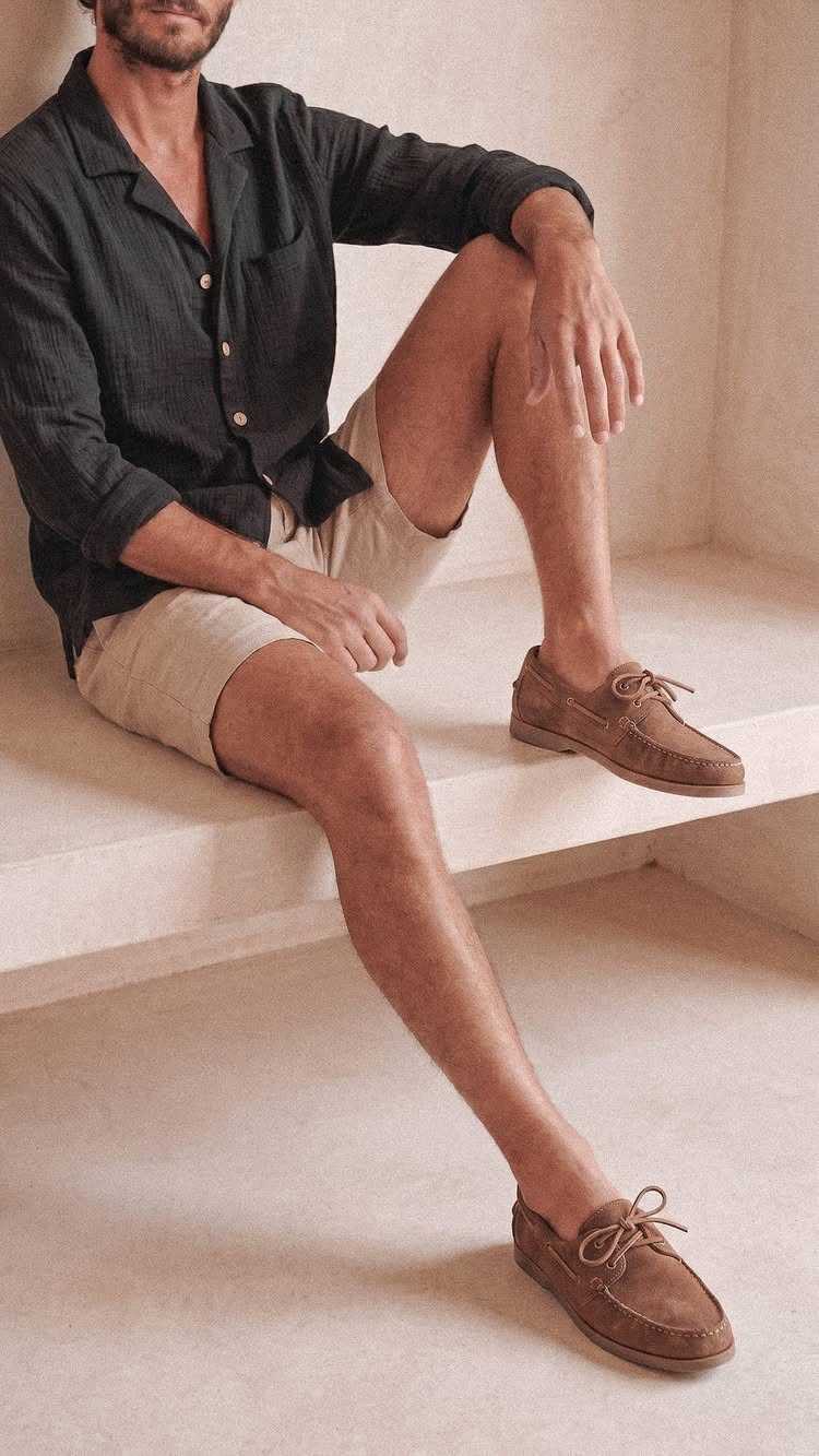 A man wearing beige shorts and brown leather boat shoes sitting on a stone ledge.