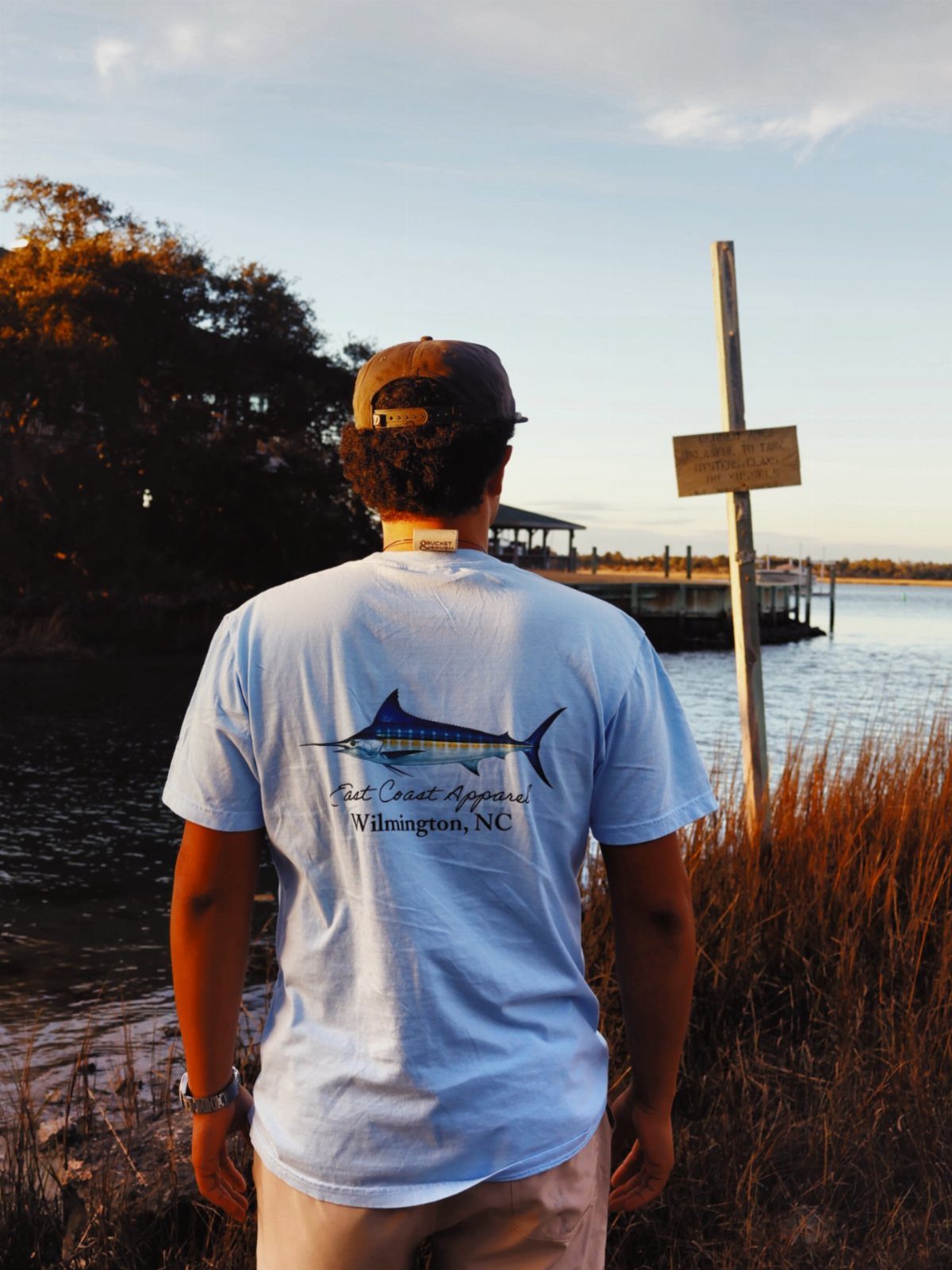 Man wearing a light blue graphic t-shirt standing by the water at sunset.