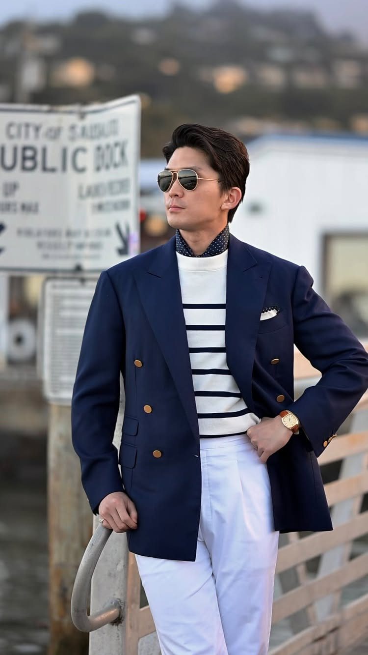 Man in a navy blazer and striped sweater posing on a wooden dock near a sign.