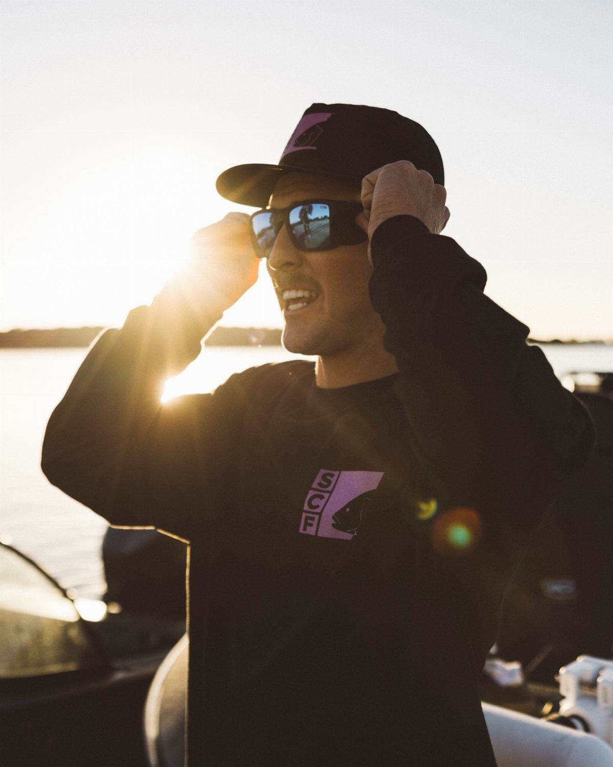 A man in a black sweatshirt and cap adjusts his dark sunglasses against a sunset.