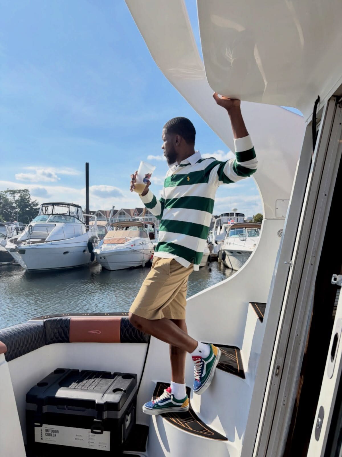 A man in a striped shirt and tan shorts posing on a boat in a marina.