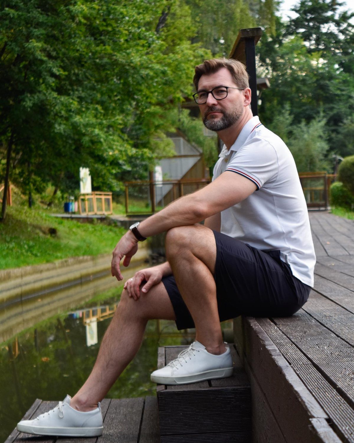 A man in a white polo and dark shorts sitting on a wooden dock.